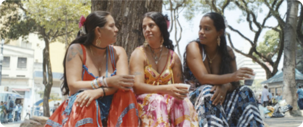 Fotografia de três mulheres sentadas lado a lado em uma praça, conversando. Elas têm cabelos longos e usam diversos adornos e roupas coloridas.