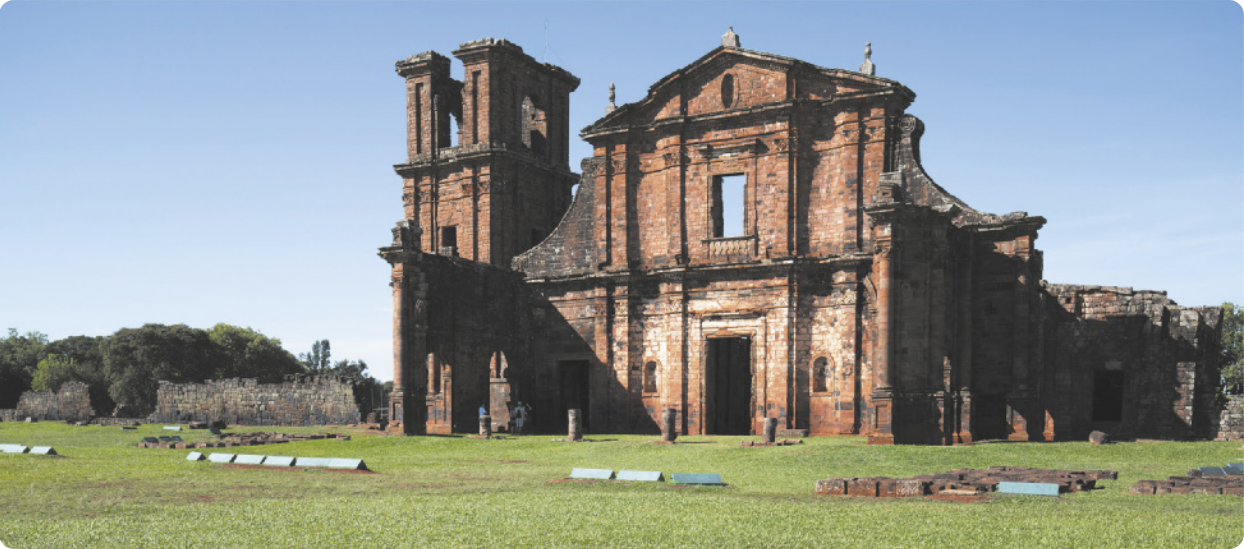Fotografia de ruínas de uma igreja com uma torre ao lado da fachada. Em frente à igreja há um gramado e há árvores ao fundo.
