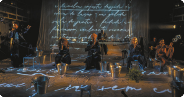 Fotografia de pessoas em um palco com a projeção de um texto manuscrito sobre o pano de fundo e o chão do palco. Há pessoas sentadas em cadeiras e um homem está em pé, falando com um microfone na mão, gesticulando com a outra mão.