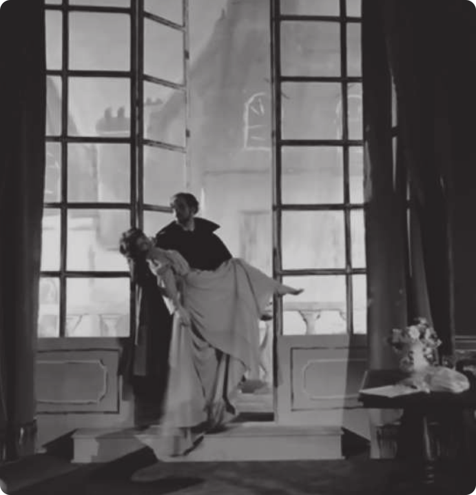 Fotografia em preto e branco de um ator carregando no colo uma atriz com um vestido longo, em frente a uma varanda com portas de vidro, em um cenário teatral.