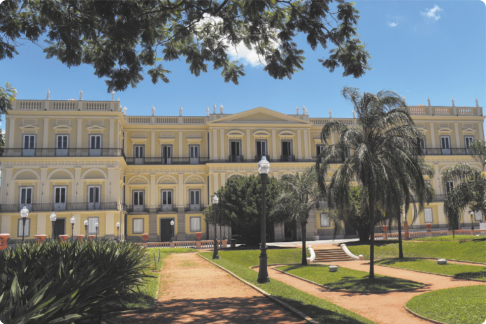 Fotografia de um edifício de arquitetura neoclássica com três andares e uma fachada larga e imponente. Em frente ao edifício, há um jardim com canteiros de grama, postes e palmeiras.
