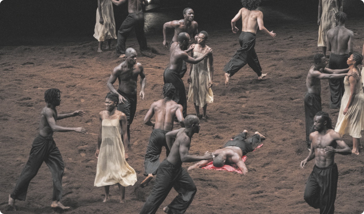 Fotografia de bailarinos e bailarinas dançando em um palco coberto de terra. Alguns bailarinos estão correndo, as bailarinas estão paradas em pé, e há um homem deitado de bruços no chão.