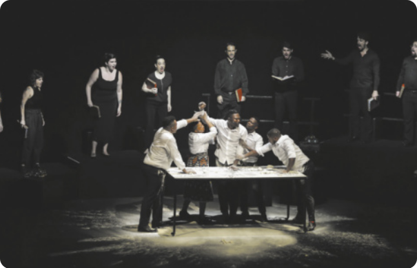Fotografia de um grupo de artistas em um palco. À frente estão artistas negros vestidos de branco, contracenando diante de uma mesa; ao fundo, estão artistas brancos vestidos de preto cantando, segurando cadernos.