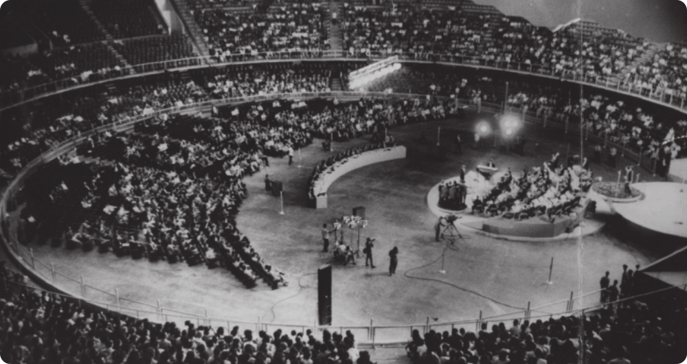 Fotografia em preto e branco de um palco circular na arena central de um estádio esportivo, com grandes arquibancadas lotadas de pessoas. Uma longa bancada semicircular de jurados está diante do palco, onde há artistas e uma orquestra.