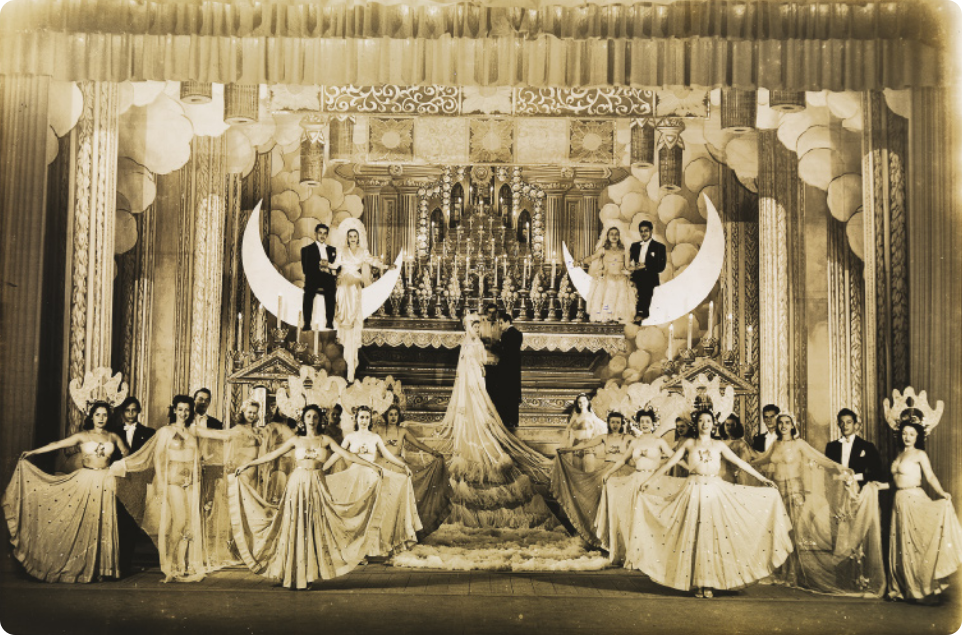 Fotografia em preto e branco de um grupo de artistas em um palco. Há um grande grupo de mulheres de vestido, segurando as saias com os braços abertos ao lado do corpo; um altar de igreja, ao fundo, com um homem e uma mulher com um vestido de noiva muito longo, que se arrasta pelo palco; e dois casais sentados em luas minguantes no alto do cenário, à esquerda e à direita do altar.