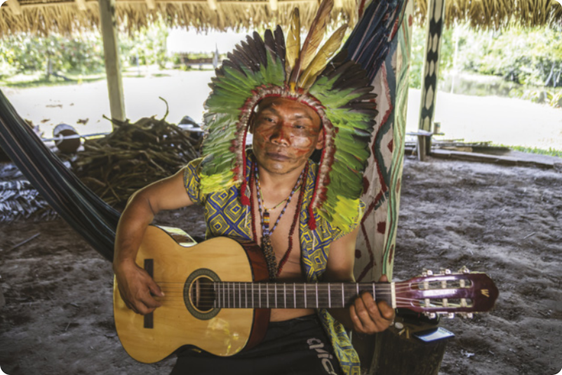 Fotografia de um homem indígena sentado tocando um violão, usando um cocar de penas e um colete com grafismos indígenas coloridos. Ele está embaixo de um telhado de palha em uma aldeia.