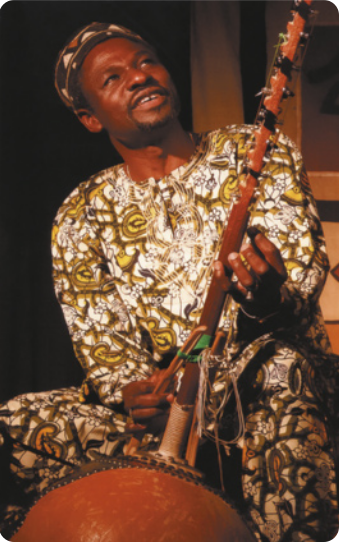 Fotografia de Toumani Kouyaté. Ele está sentado, com roupas estampadas e um chapéu, tocando um instrumento de cordas com uma cabaça e um braço de madeira em que estão esticadas as cordas do instrumento.