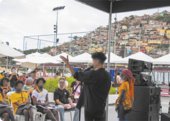 Fotografia de um homem em um palco, falando com um microfone na mão, gesticulando com a outra mão aberta e erguida à altura da cabeça.