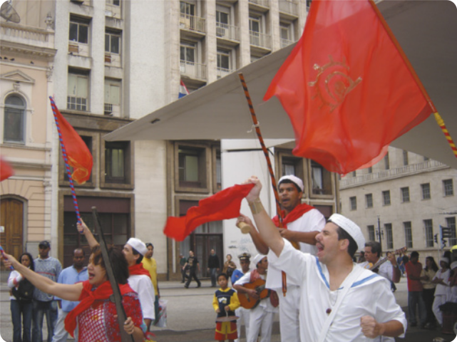 Fotografia de um grupo de atores se apresentando em uma praça. Alguns deles estão usando trajes de marinheiro e brandindo bandeiras vermelhas; há uma atriz segurando uma espingarda, com um lenço vermelho no pescoço; e há homens tocando violões ao fundo.
