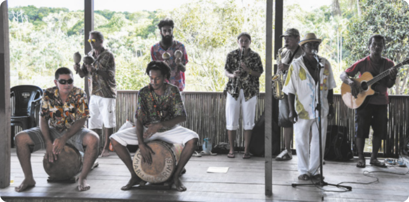 Fotografia de um grupo de homens com camisas coloridas tocando instrumentos musicais em um palco. Dois deles tocam tambores grandes, sentados sobre eles; dois deles tocam chocalhos; outros dois tocando saxofone e clarinete; um deles toca violão; e outro está cantando diante de um microfone.