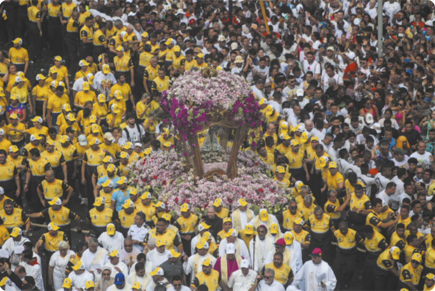 Fotografia de uma procissão com um grande grupo de pessoas caminhando uniformizadas, com bonés e camisetas amarelas, carregando uma berlinda decorada com flores, com uma imagem de Nossa Senhora no centro da berlinda. Ao lado do grupo de pessoas uniformizadas caminhando há uma multidão de pessoas assistindo à procissão, separadas da procissão por uma linha de homens vestidos de branco atuando como um cordão de isolamento.