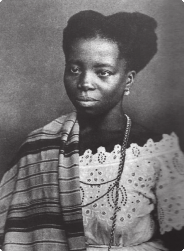 Fotografia em preto e branco de Hilária Batista de Almeida. Ela é uma mulher com brincos, colares e um vestido, usando um manto de tecido listrado sobre o ombro direito.