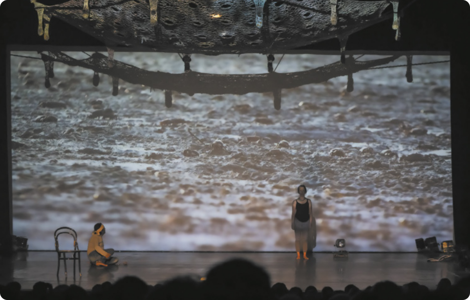 Fotografia de Ailton Krenak e Andreia Duarte em um palco, com a imagem de um solo de areia com muitos caranguejos projetada em uma tela ao fundo do palco. Ailton Krenak está sentado no chão, ao lado de uma cadeira, usando uma faixa no cabelo; e Andreia Duarte está em pé do outro lado do palco, descalça, voltada para a plateia.