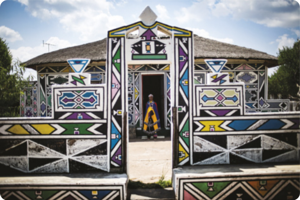 Fotografia de uma casa com telhado de palha cuja fachada e muros são pintadas com motivos geométricos abstratos de cores vivas, com formas triangulares, setas, losangos, retângulos, entre outras formas.