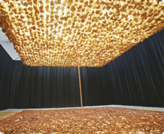 Fotografia de instalação artística com uma grande quantidade de moedas e hóstias espalhadas em um quadrado no chão delimitado por pedras de pavimento, em um ambiente fechado por cortinas de tecido preto em volta das pedras. No alto da instalação estão pendurados muitos ossos de boi, que formam uma espécie de teto sobre a instalação.