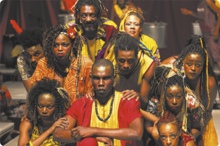 Fotografia de um grupo de homens e mulheres sentados e agachados, com expressão séria, usando adereços, penteados de tranças e roupas com estampas de inspiração africana.
