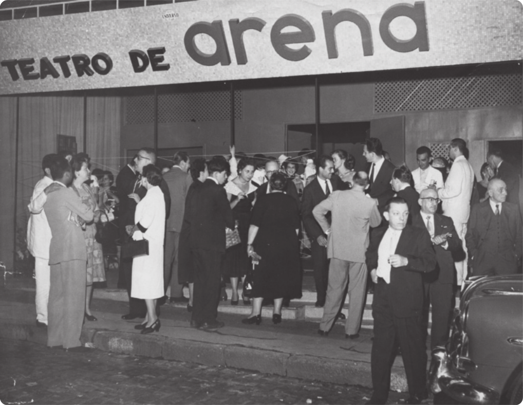 Fotografia em preto e branco de um grupo de pessoas conversando em uma calçada em frente à entrada de um teatro, usando ternos e vestidos.