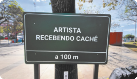 Fotografia de uma placa de trânsito em uma via pública com o seguinte texto: 'Artista recebendo cachê a 100 metros'.