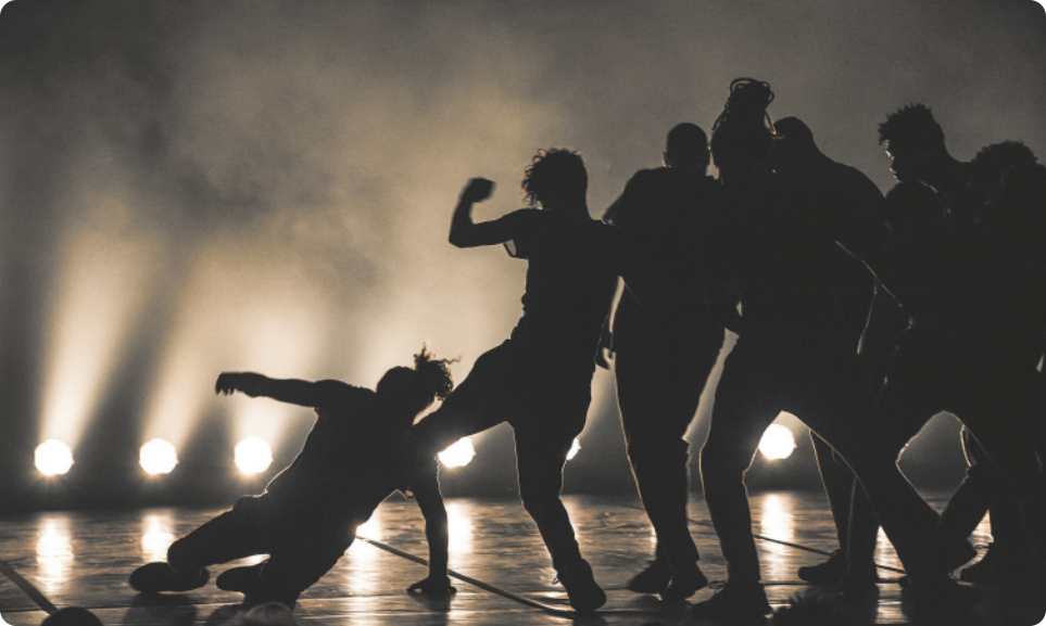 Fotografia de um grupo de pessoas em um palco dançando à contraluz, de modo que só se vê as silhuetas delas, com fumaça cênica e holofotes ao fundo. Há um homem executando um passo de dança no chão, e os outros estão em pé, com os corpos muito próximos e em poses diversas.