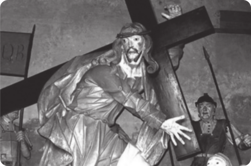 Fotografia em preto e branco de uma escultura representando Jesus Cristo carregando um crucifixo de madeira nas costas. Ao fundo há uma escultura representando um arauto com trajes militares, segurando uma lança.