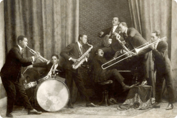 Fotografia em preto e branco de um grupo de músicos vestidos com ternos, tocando instrumentos diversos como piano, banjo, trombone, bateria, saxofone e trompete. Ao centro, em pé, está Pixinguinha, um dos dois saxofonistas do grupo.