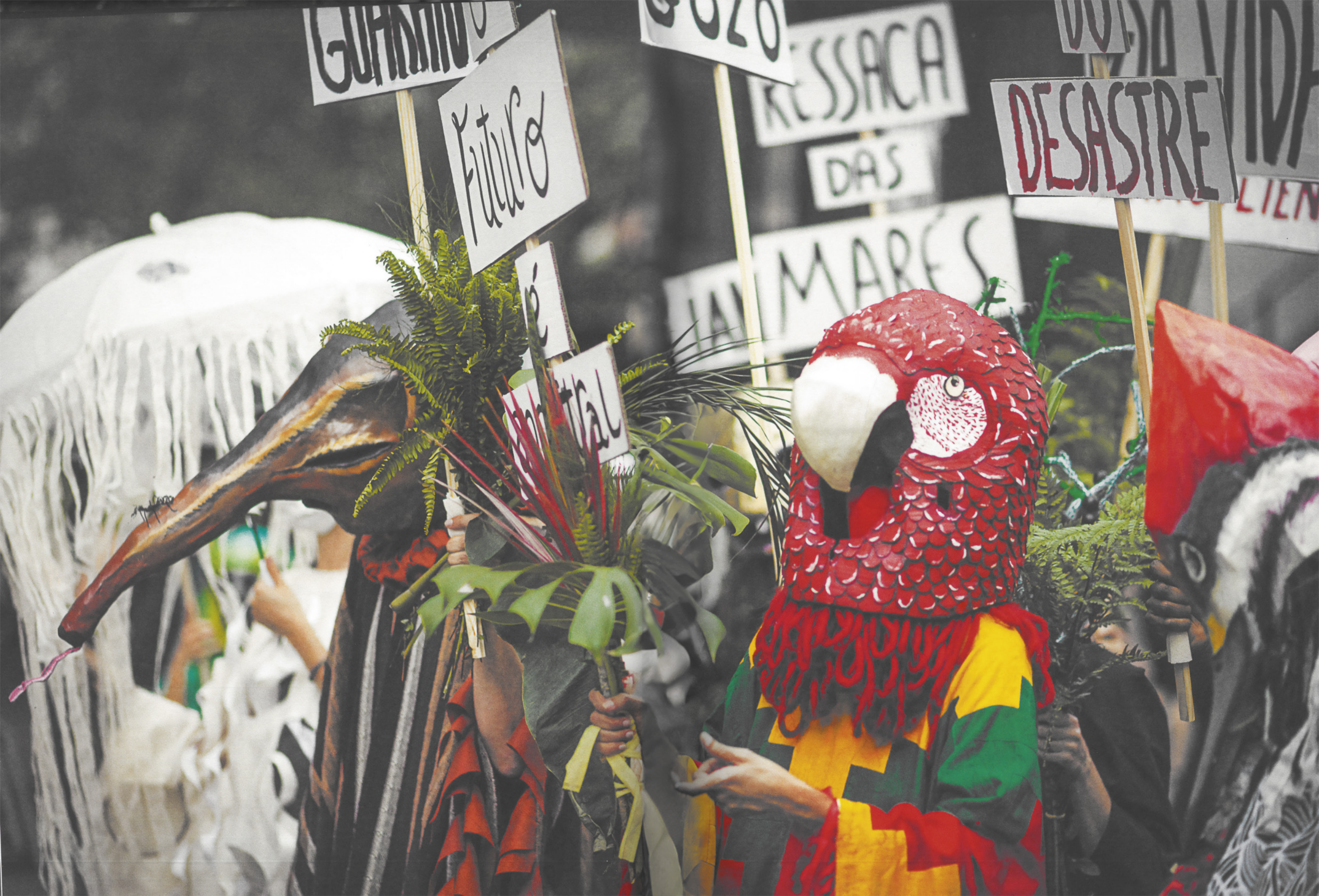 Fotografia de um grupo de pessoas usando fantasias com cabeças de animais como tamanduá e arara, enfeitadas com folhas e plantas, carregando placas em que se lê palavras e mensagens como 'desastre', 'ressaca das marés', e 'o futuro é ancestral'.
