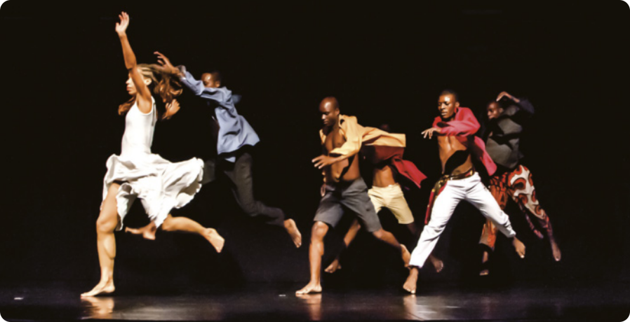 Fotografia de um grupo de dançarinos e dançarinas em um palco, correndo e saltando no ar, em fila.