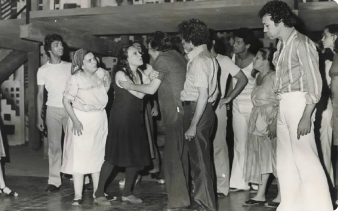 Fotografia em preto e branco de um grupo de atores reunidos em um palco, encenando uma discussão, protagonizada por uma mulher e um homem ao centro, que estão segurando os braços um do outro. As pessoas em volta se dirigem a eles e participam da discussão.