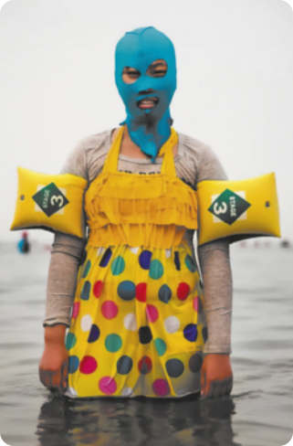 Fotografia de uma mulher com as pernas submersas na água do mar, usando um vestido com estampa de círculos coloridos e boias nos braços. Ela está usando uma máscara de tecido elástico que cobre a cabeça, o rosto e o pescoço dela, com orifícios para os olhos, boca e nariz.