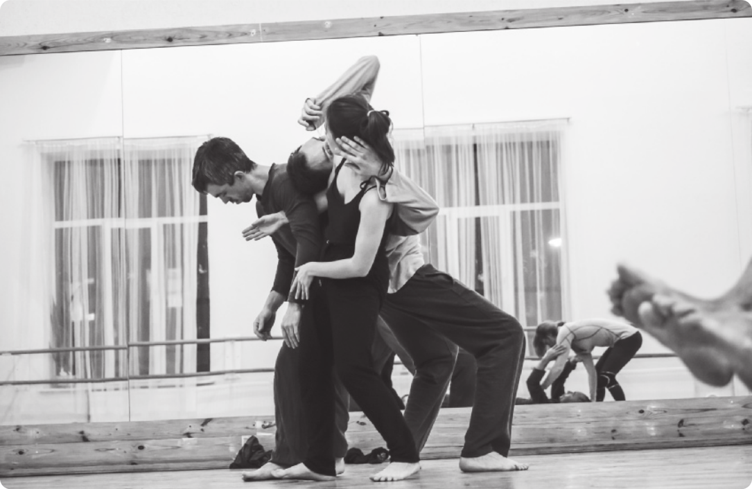 Fotografia em preto e branco de dois homens e uma mulher em uma sala de dança realizando um movimento em que um dos homens inclina o corpo para trás, deitando-se sobre as costas do homem e da mulher que estão com ele; estes, por sua vez, estão de braços dados e com as pernas paralelas e flexionadas, para dividir a carga do corpo do homem que se inclina sobre eles.