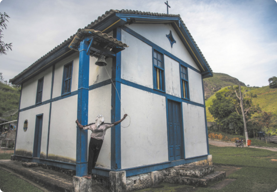 Fotografia de uma pequena igreja de arquitetura do período colonial, com um sino instalado ao lado da fachada, em uma paisagem de morros. Há um homem puxando uma corda que balança o sino.