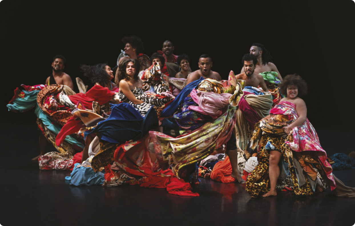 Fotografia de um grupo de homens e mulheres dançando em um palco, vestidos com muitos tecidos, mantas e cobertores com estampas diversas enrolados no corpo.