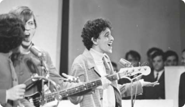 Fotografia em preto e branco de Caetano Veloso: um homem de cabelos cacheados curtos, usando um paletó e uma blusa de gola alta, cantando com os braços em um palco, diante de um microfone. Ao lado dele estão dois músicos cantando diante de um microfone, um deles tocando contrabaixo elétrico, e o outro tocando pandeiro.