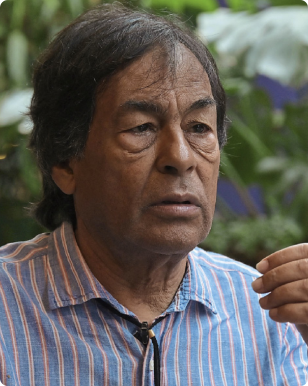 Fotografia de Ailton Krenak. Ele é um homem indígena, de cabelos castanhos, lisos e curtos, com linhas de expressão visíveis abaixo dos olhos. Veste uma camisa listrada e usa um cordão ao redor da gola. Está com a boca entreaberta e faz um gesto com a mão, como se estivesse conversando.