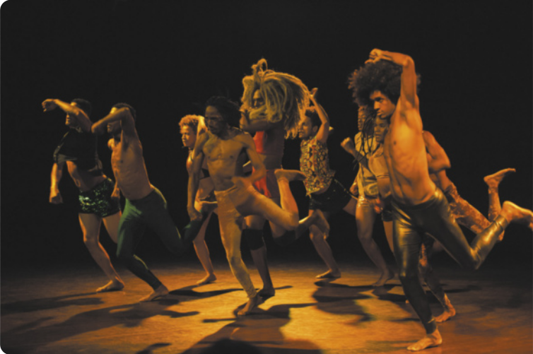 Fotografia de um grupo de homens e mulheres dançando em coreografia em um palco, dispostos em duas fileiras. Eles estão realizando um movimento em que dão passadas com o corpo inclinado para a frente, levantando a perna esquerda para trás com o joelho flexionado, e erguendo o braço esquerdo, com o cotovelo flexionado, acima da cabeça.