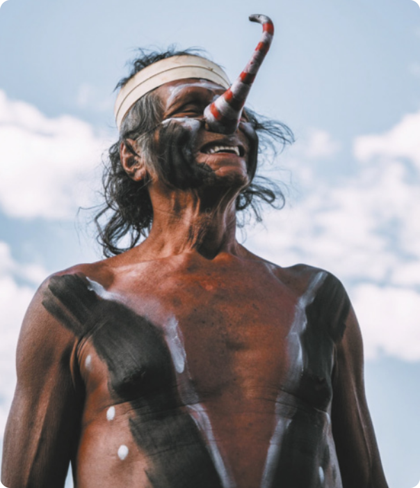Fotografia de Ismael Aprac Krahô. Ele é um homem indígena com cabelos grisalhos e pinturas corporais no tronco e no rosto. Ele está usando uma faixa no cabelo e uma prótese de nariz de formato alongado, com uma curva para cima, pintado com listras vermelhas.