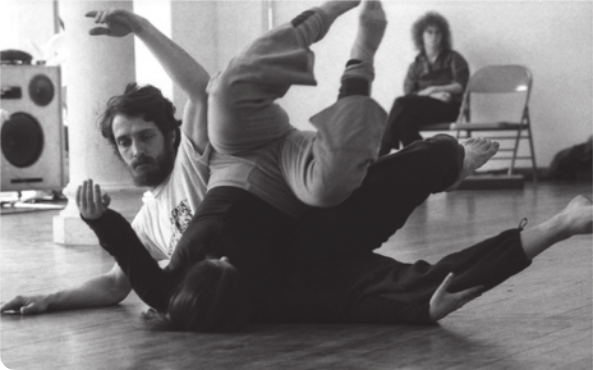 Fotografia em preto e branco de Steve Paxton. Ele é um homem com barba e cabelos curtos ondulados, deitado de lado no chão com um braço erguido para o alto e com os joelhos flexionados, erguendo os pés para cima. Uma mulher está dançando com ele, deitada de cabeça para baixo com as costas apoiadas sobre o tronco do dançarino, flexionando as pernas, com as pontas dos pés apontando para cima, e os braços estendidos paralelos ao chão.