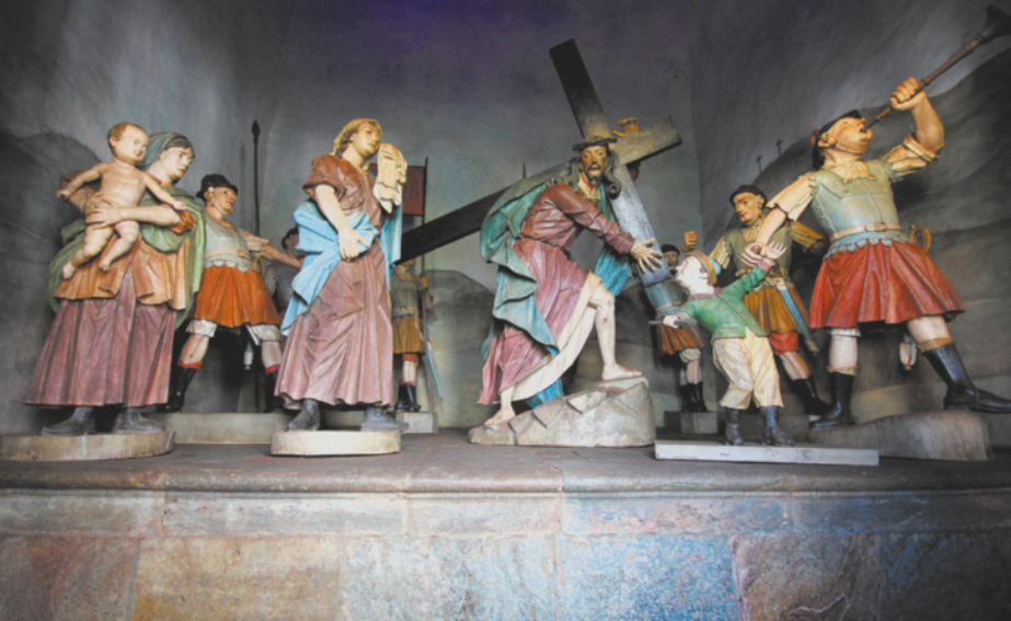 Fotografia de um conjunto de esculturas representando figuras diversas caminhando em torno da imagem de Jesus Cristo curvado, carregando uma cruz, subindo um pedestal em forma de rampa. Em volta de Jesus há um menino de chapéu que se dirige a ele, porém é puxado pela mão por um militar; uma mulher com um lenço na cabeça, carregando um bebê; um homem de túnica, com um lenço na cabeça, carregando um pedaço de tecido; e militares de capacete e armaduras, alguns deles carregando lanças, e um deles tocando uma trombeta.