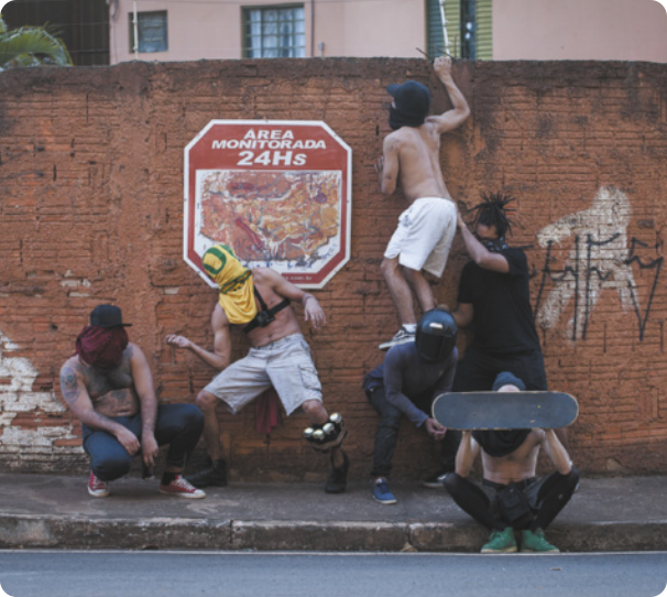 Fotografia de um grupo de seis homens com os rostos cobertos por camisas, panos, lenços, um skate e um capacete. Eles estão em uma calçada, sentados, agachados e em pé, e um deles está em pé sobre as costas de outro, escalando o muro atrás da calçada.