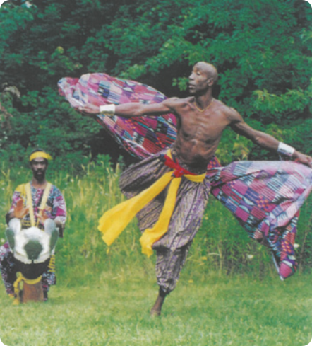 Fotografia de um homem dançando em um gramado, com a mata ao fundo. Ele está com os braços abertos, sem camisa, usando uma calça larga com uma faixa colorida, segurando um tecido estampado com grafismos abstratos africanos. Atrás dele há um homem sentado tocando um tambor.