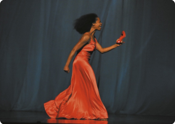 Fotografia de Regina advento. Ela é uma bailarina com cabelos longos e cacheados, usa vestido e está andando em um palco, segurando um sapato de salto alto.