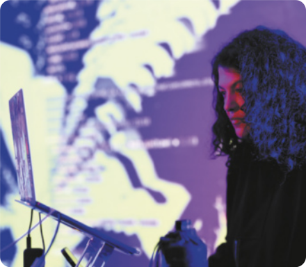 Fotografia de Alexandra Cárdenas. Ela é uma mulher com cabelos cacheados e longos diante de um notebook, com uma tela projetando imagens luminosas ao fundo.