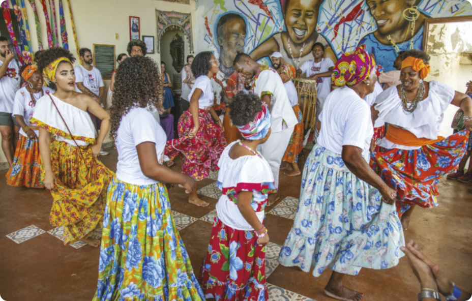 Fotografia de um grupo de mulheres dançando em um salão, com saias de estampas floridas e lenços na cabeça. Ao fundo há um grupo de homens tocando instrumentos de percussão.