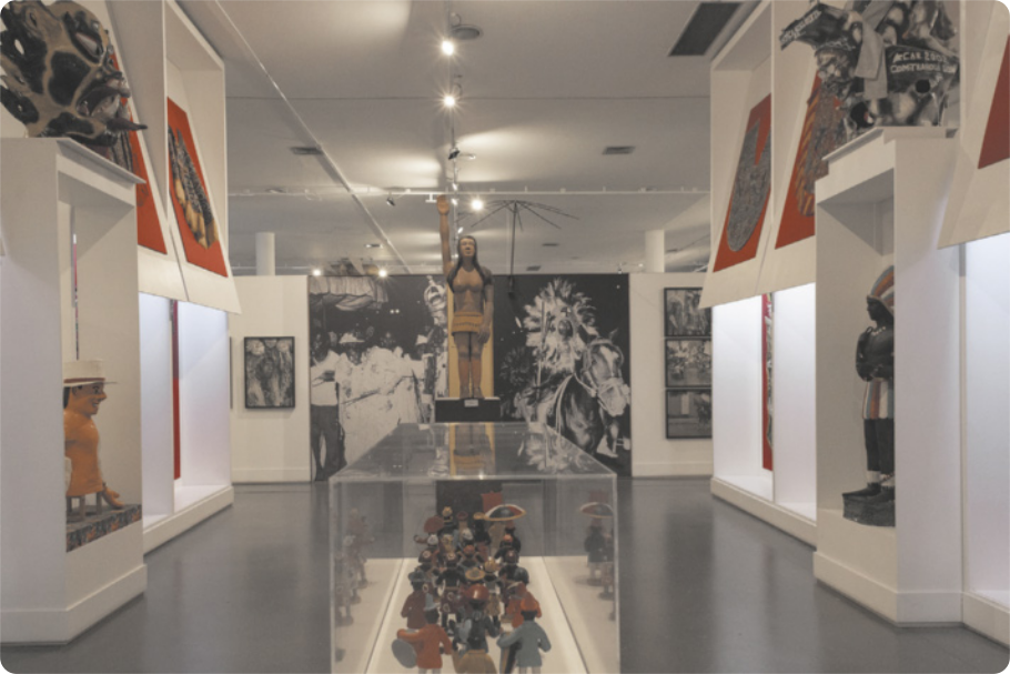 Fotografia de sala expositiva com diversas esculturas de figuras humanas do universo cultural popular afrodescendente, quadros com fotografias nas paredes e outros objetos escultóricos.