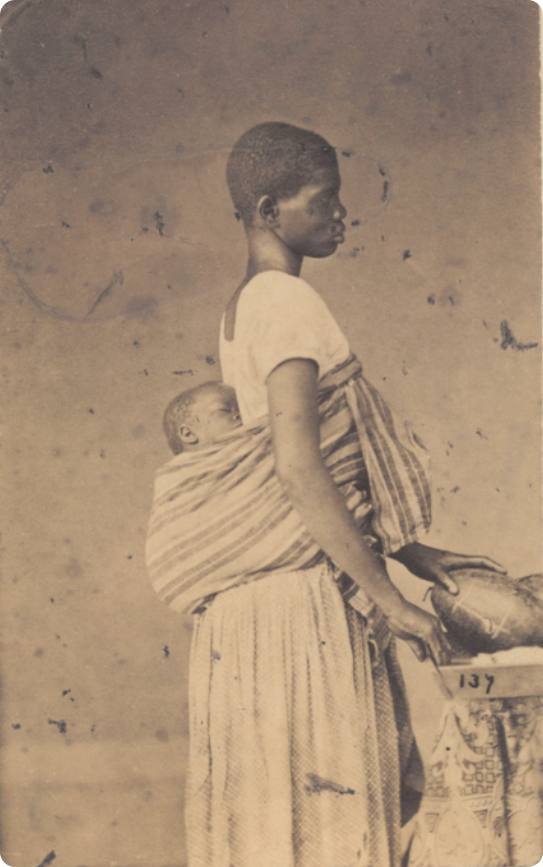 Fotografia em preto e branco de uma mulher escravizada de vestido, com cabelos curtos, carregando um bebê nas costas, enrolado em uma faixa de tecido. Ela está em pé, de perfil, diante de uma mesa com frutas.