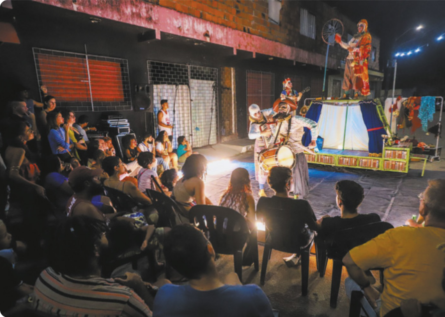 Fotografia de um grupo de atores se apresentando ao ar livre em um a via pública, diante de uma plateia de pessoas sentadas em cadeiras. Os artistas estão fantasiados, e tocam instrumentos musicais, como violão, sanfona e um tambor. Ao fundo, há um pequeno palco sobre o qual está em pé um dos atores.