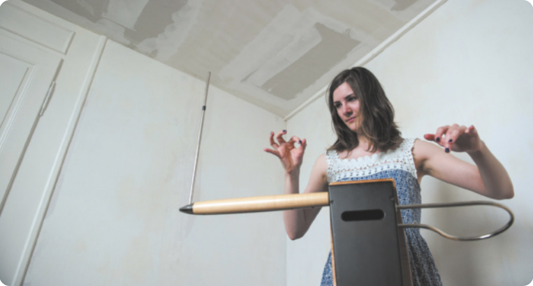 Fotografia de Carolina Eyck: uma mulher de cabelos longos e lisos, usando um vestido. Ela está tocando um teremim, gesticulando com as mãos no ar em frente ao corpo, diante do instrumento.