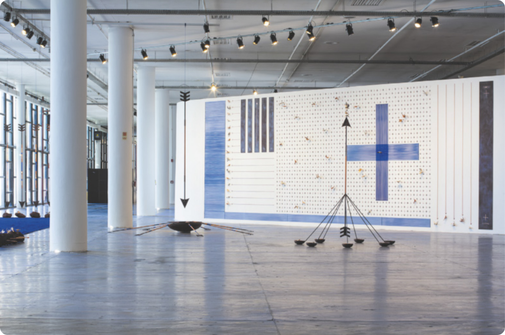 Fotografia de um espaço expositivo com instalação artística. A instalação consiste em um grande painel com objetos metálicos pendurados por cordas, formando uma composição de diversas formas abstratas: linhas e faixas verticais e horizontais, cruzes, e uma trama de pontos justapostos. Em frente ao painel, há duas esculturas com flechas de arame: uma delas apontada para cima, estruturada em hastes de metal apoiadas sobre potes de ferro; e a outra para baixo pendurada no teto apontando para uma grande tigela de ferro no chão.