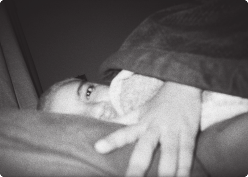 Fotografia em preto e branco de uma pessoa deitada em uma cama, com o corpo coberto por uma manta. Ela está com uma das mãos e parte do rosto aparentes ao centro da imagem, iluminados, e o resto da composição da imagem está obscurecido, formando planos, eixos e linhas que favorecem visualmente a porção central da imagem.