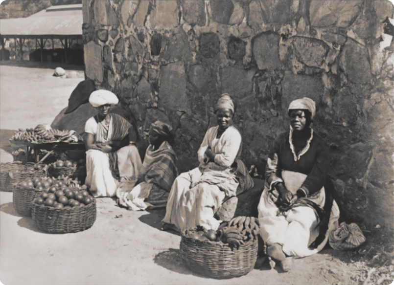 Fotografia de quatro mulheres escravizadas usando lenços na cabeça, sentadas ao lado de um muro de pedras. Ao lado delas há diversos cestos de palha trançada com frutas.